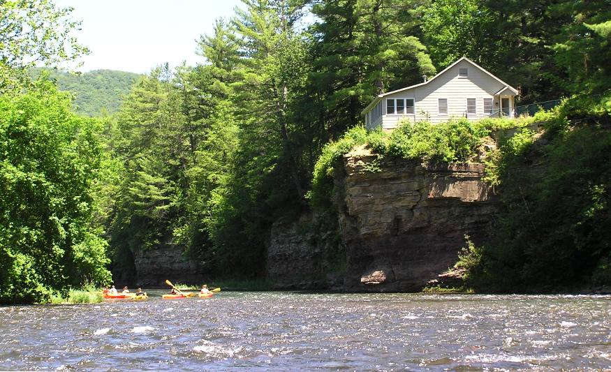 Kayakers on riffles near a steep, rocky cliff with a house on it