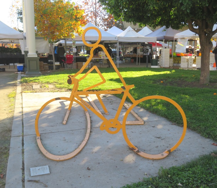 Yellow metal sculture of bicyclist from different angle shows him upright