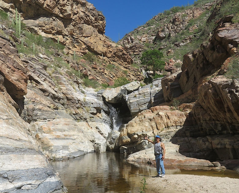 Me with arm lifted as if presenting the waterfall while standing on a small beach