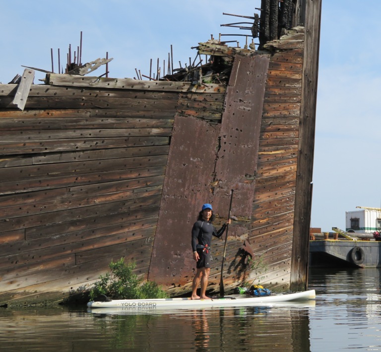 Me on SUP with shipwreck behind pieced together with lots of wooden dowels