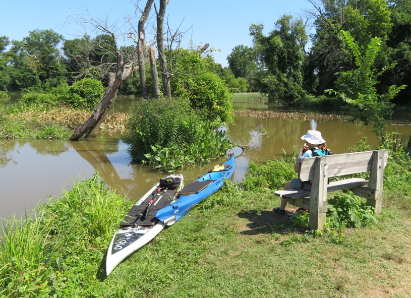 SUP and kayak on shore near bench where Norma is sitting