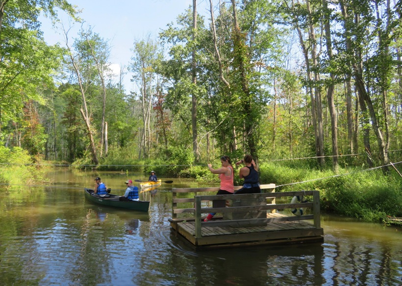 People pulling ropes to move a wooden raft across the creek