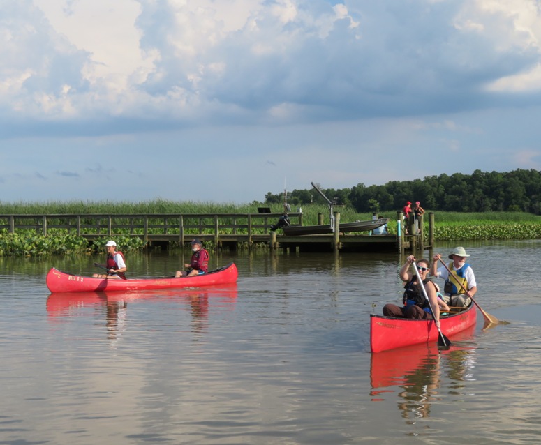 People in canoes paddling upstream with the river pier in the background