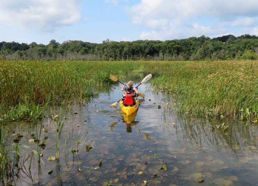 Aft view of Beate and Gerhard kayaking in narrow, grass-lined waterway